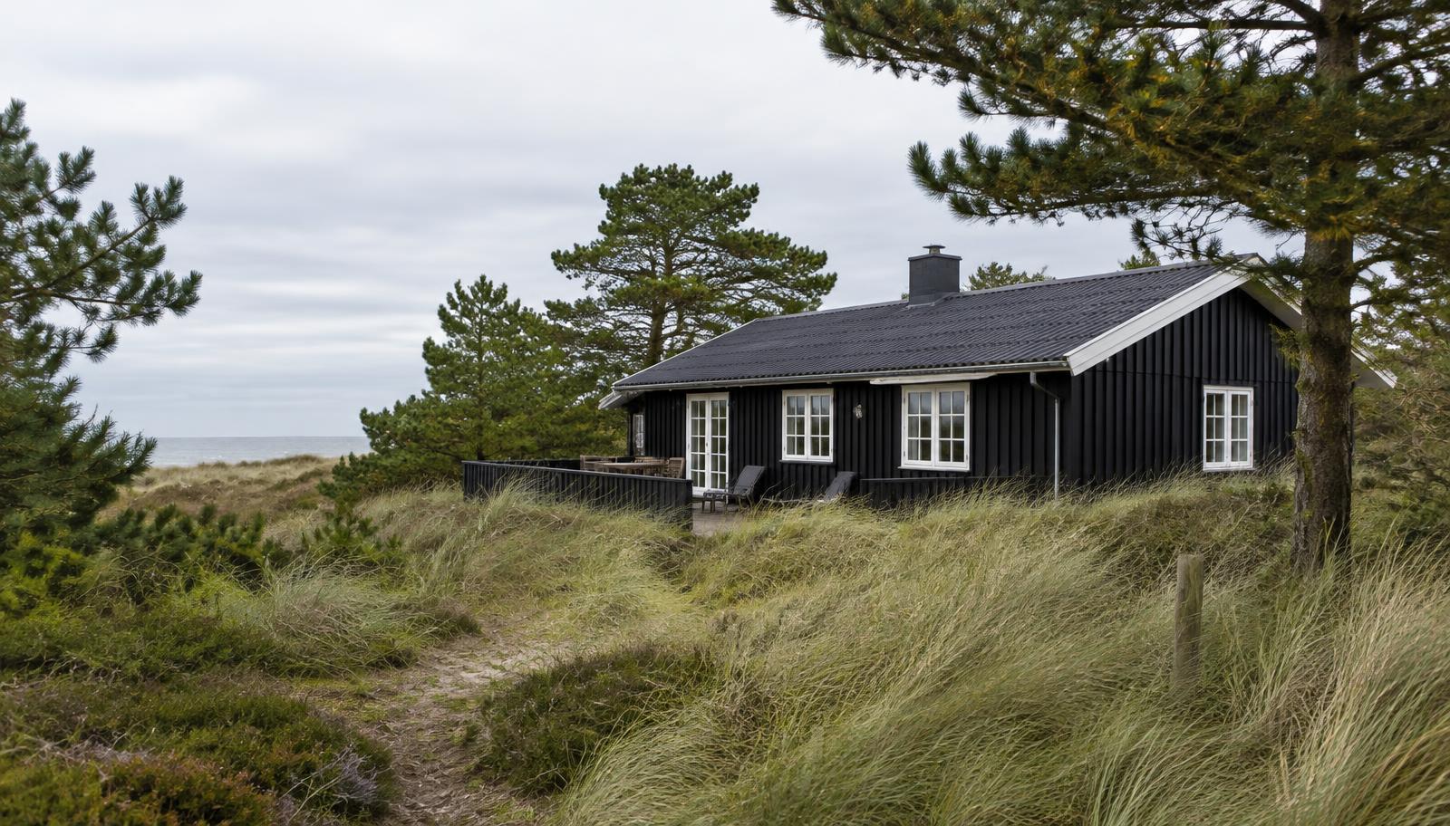 Sortmalet træsommerhus med hvide vinduer mellem klitter, marehalm og fyrretræer ved den danske kyst