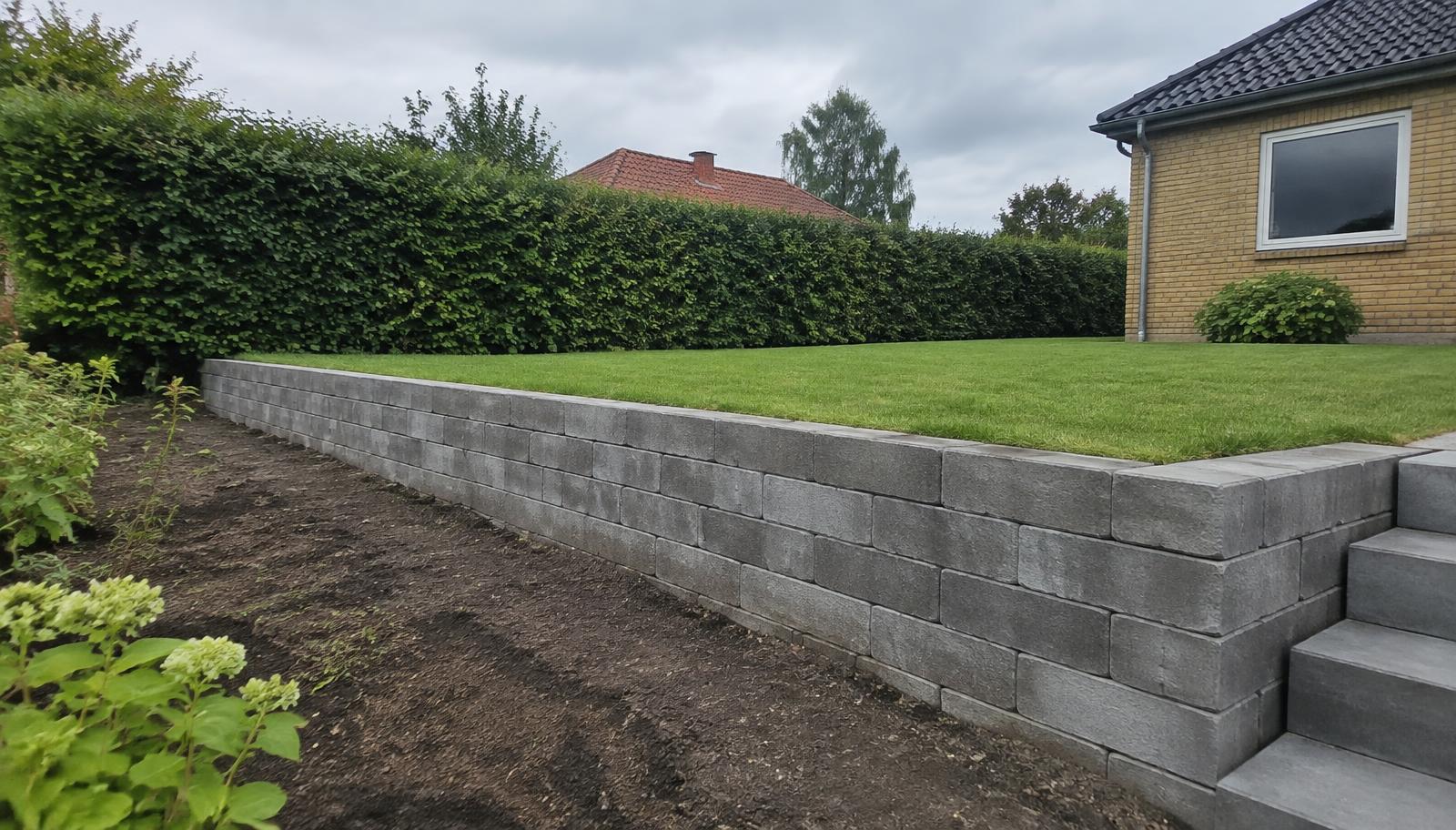 Støttemur i grå betonblokke i skrånende dansk have med plan græsplæne